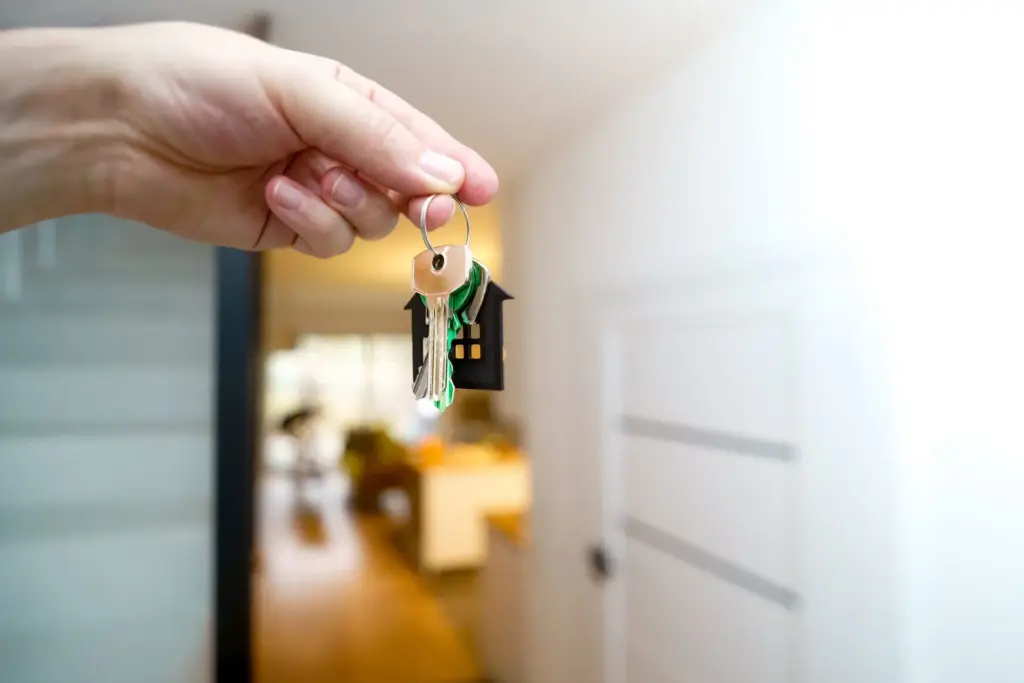 A person’s hand displaying a key, with a house visible behind, illustrating the idea of unlocking a new home.